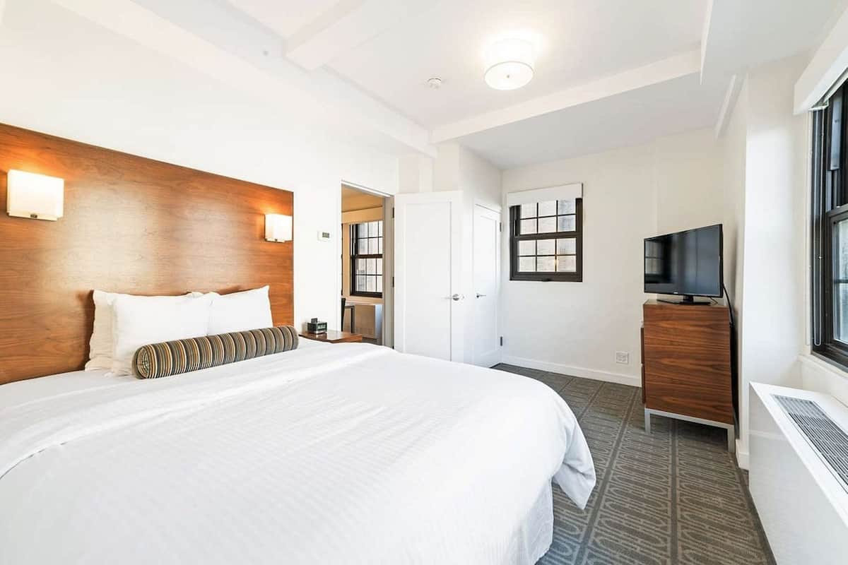 A hotel room with a large bed, a wooden headboard, a TV on a wooden cabinet, and a window with blinds.