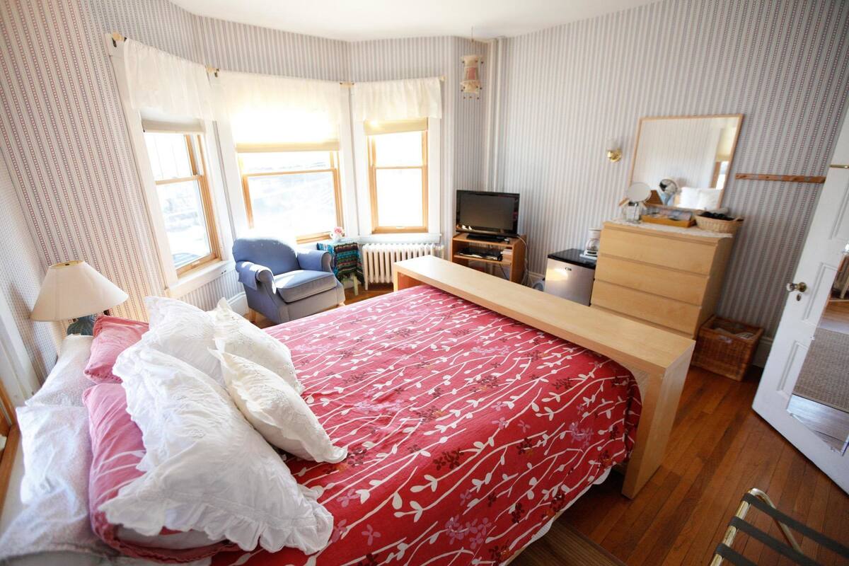 A bedroom with a bed, a desk, a chair, a dresser, a television, and a window.