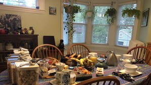 A dining table set with breakfast items, including bread, fruit, and beverages, in a room with wooden chairs and a window with plants. - A Village Bed and Breakfast (Newton)