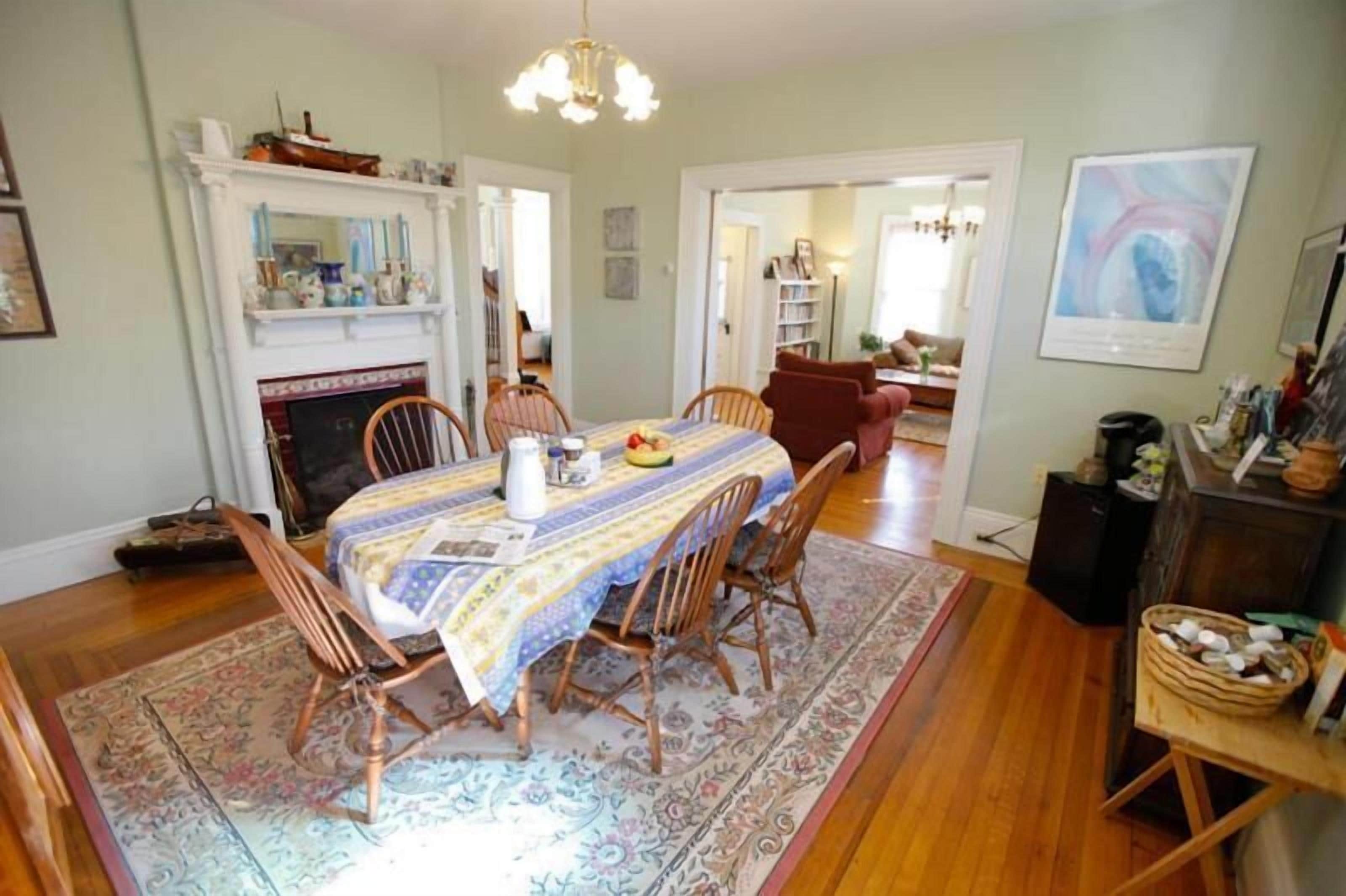 A dining area with a table set for four, surrounded by wooden chairs. A patterned rug covers the floor, and a fireplace is visible in the background.