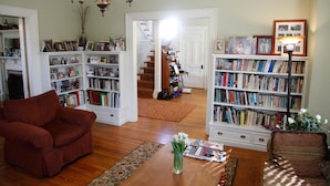 A living room with a red armchair, a wooden coffee table with magazines, and two bookshelves filled with books and framed photos. - A Village Bed and Breakfast (Newton)