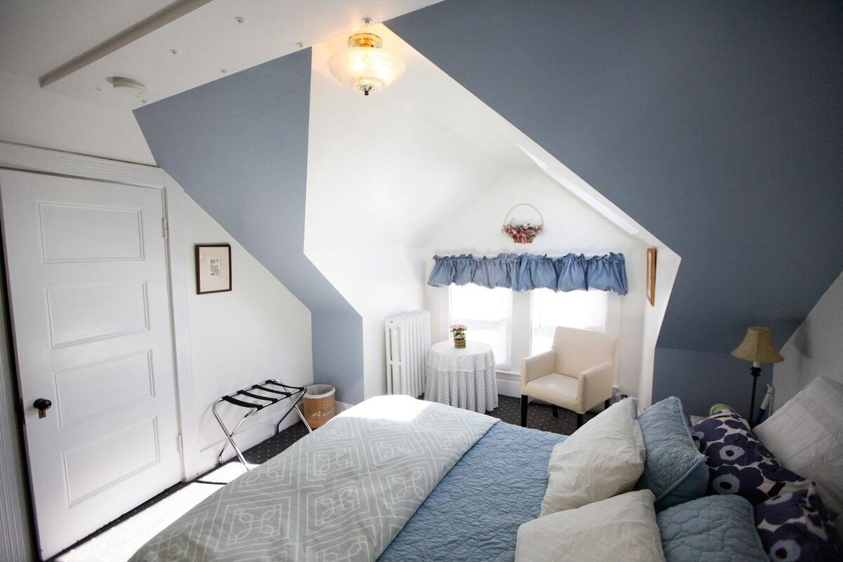 A bedroom with a sloped ceiling, a bed with patterned bedding, a chair, a small table with a vase, and a window with curtains.