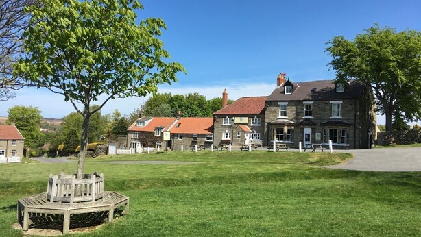 A traditional stone building with a red-tiled roof, a grassy lawn, and a wooden bench. - The Fox and Hounds (Whitby)