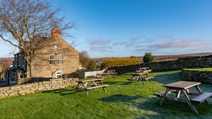 A stone building with a large tree in front, surrounded by picnic tables and a stone wall. - The Fox and Hounds (Whitby)