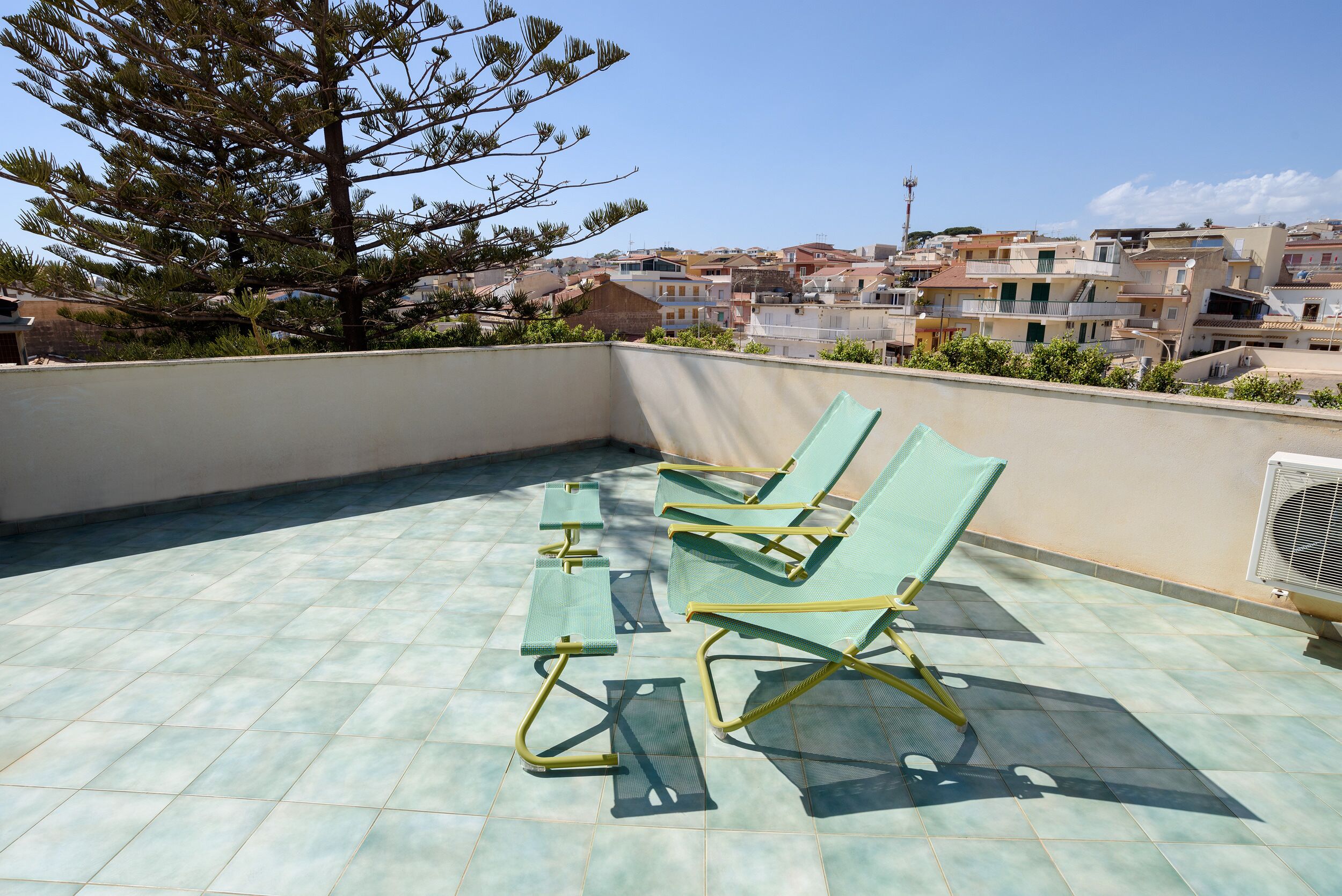 A rooftop terrace with green chairs and a table, overlooking a residential area with buildings and a tree.