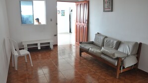 A room with a wooden floor, a white bench, a grey sofa, and a door leading to another room. - Sunset Terrace - Hostel (Cartagena)