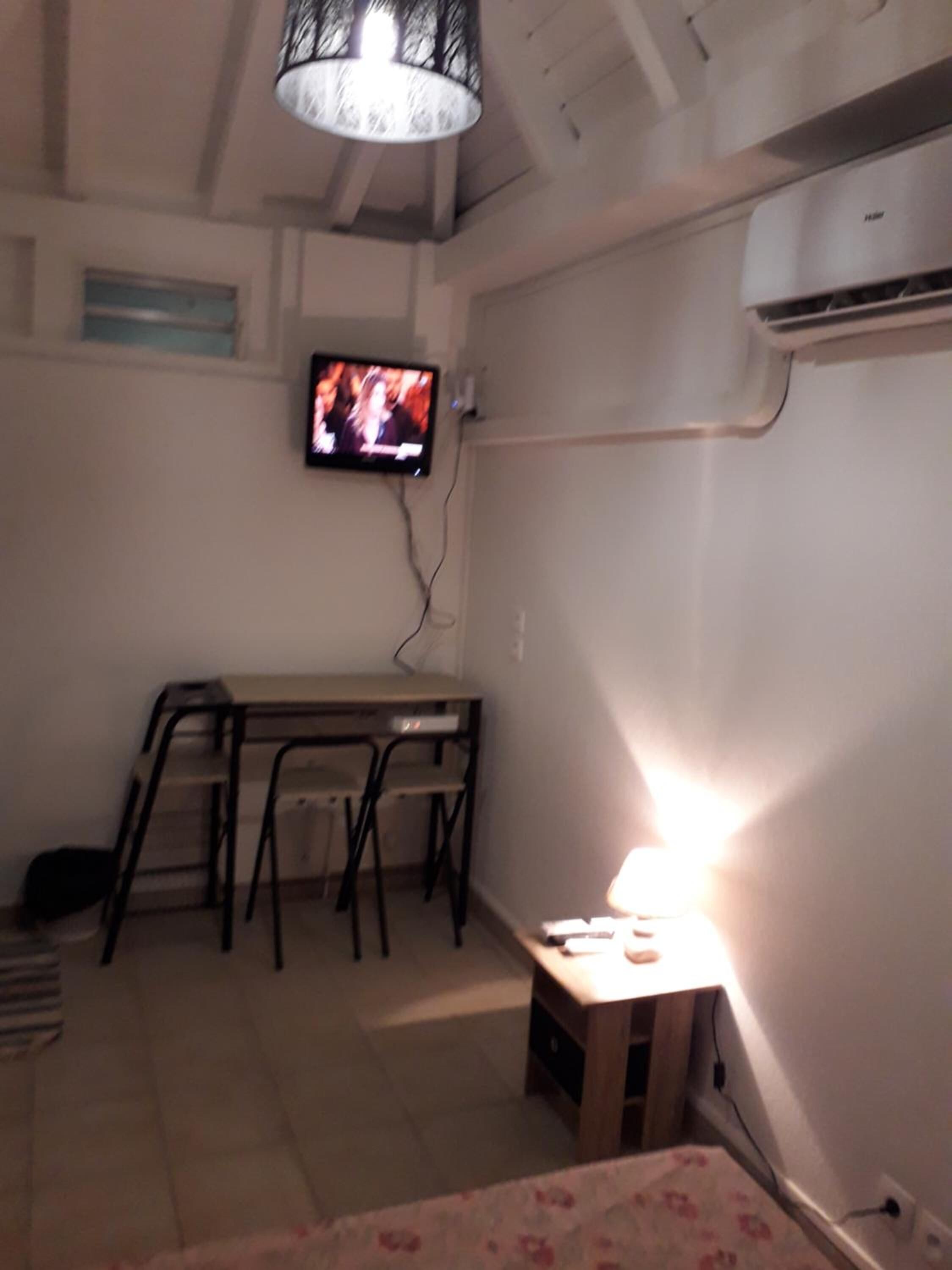 A small room with a TV mounted on the wall, a desk with chairs, and an air conditioning unit.