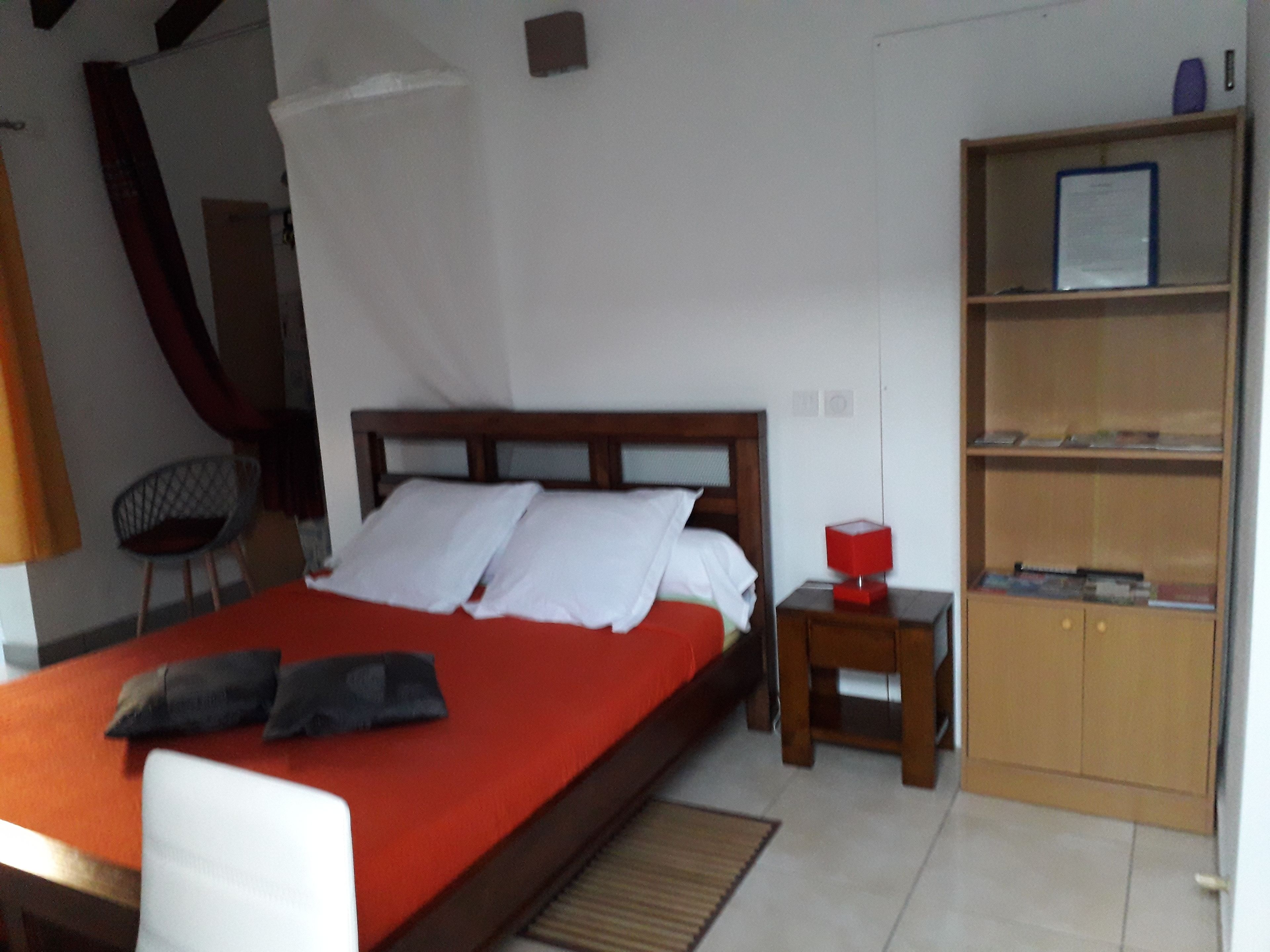 A bed with a red cover, a wooden headboard, and two pillows. There is a small wooden nightstand with a red lamp beside the bed. A bookshelf with books and a framed certificate is to the right of the bed.