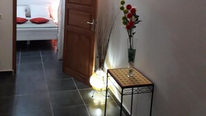 A hallway with a bed in the background, a wooden door, a small table with a vase, and a light fixture. - Le Saint Georges (Le Gosier)