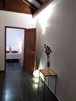 A hallway with a bed in the background, a wooden door, a small table with a vase, and a light fixture.