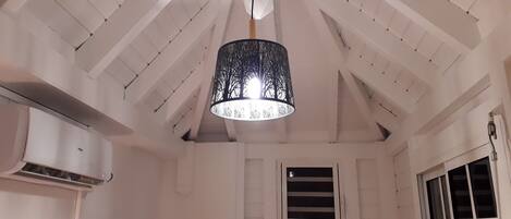 A room with a sloped ceiling, a hanging black chandelier, a white air conditioning unit, and a window with a view.