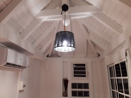 A room with a sloped ceiling, a hanging black chandelier, a white air conditioning unit, and a window with a view.