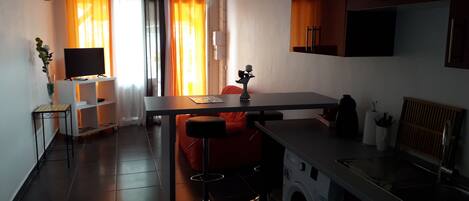 A compact kitchen with a washing machine, a dining area with a table and chairs, and a TV on a stand.