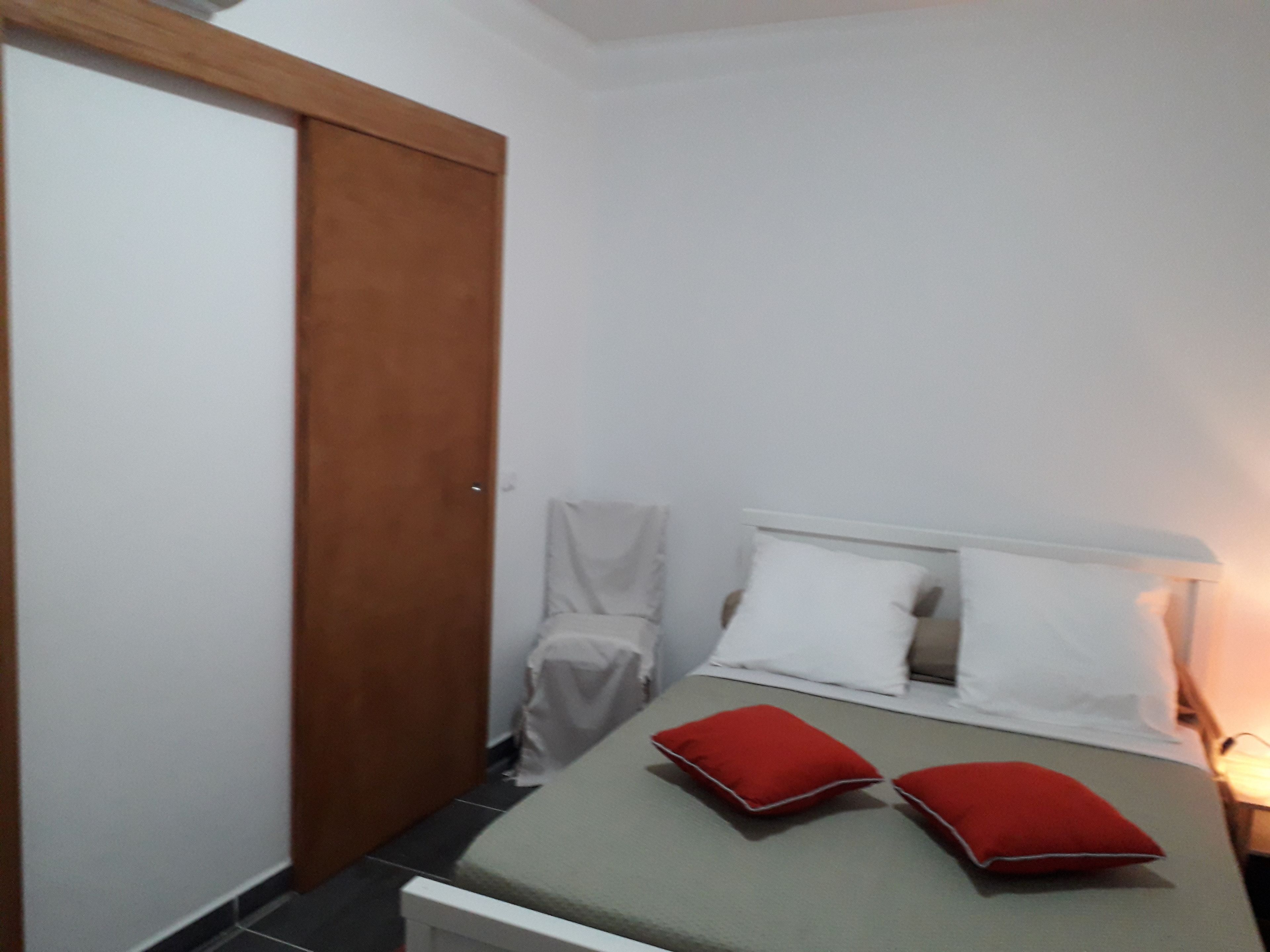 Quarto pequeno e bem organizado, com uma cama, duas almofadas, uma mesa de cabeceira e uma porta de madeira.
