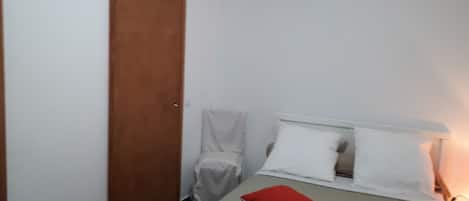 A small, neatly arranged bedroom with a bed, two pillows, a nightstand, and a wooden door.