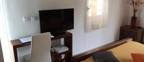 Un dormitorio con cama, escritorio con televisor, silla y un pequeño cubo de basura.