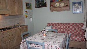A kitchen with wooden cabinets, a sink, and a stove. There is a dining table with a checkered tablecloth and chairs. - Casa Bel Prà (Val di Zoldo)