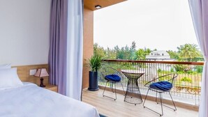 Een hotelkamer met balkon, een bed, nachtkastje, lamp en uitzicht op groen en een gebouw.