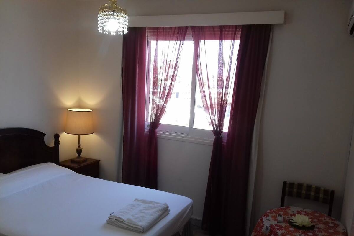 Ein Hotelzimmer mit einem Bett, einem Nachttisch mit Lampe, einem Fenster mit Vorhängen und einem kleinen Tisch mit Blumenmuster-Tischdecke.