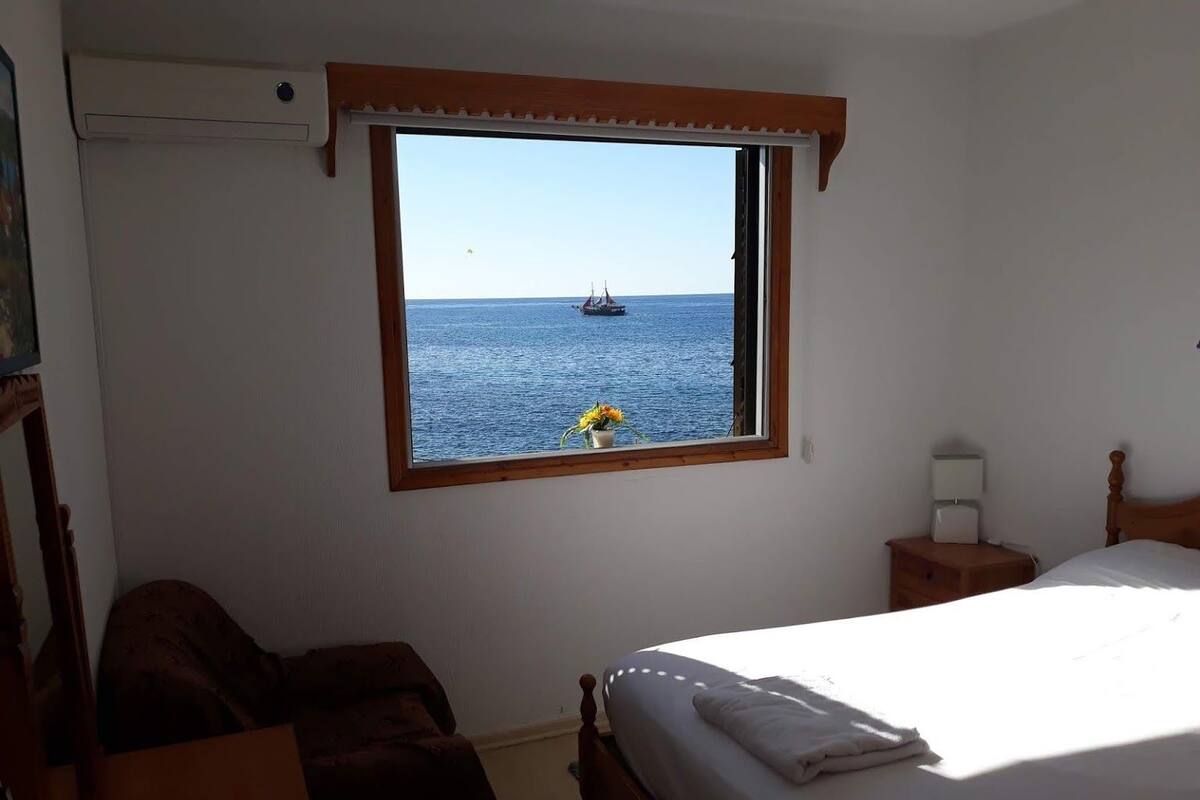 Ein Schlafzimmer mit großem Fenster und Meerblick, einem Bett, einem Nachttisch und einem Sessel.