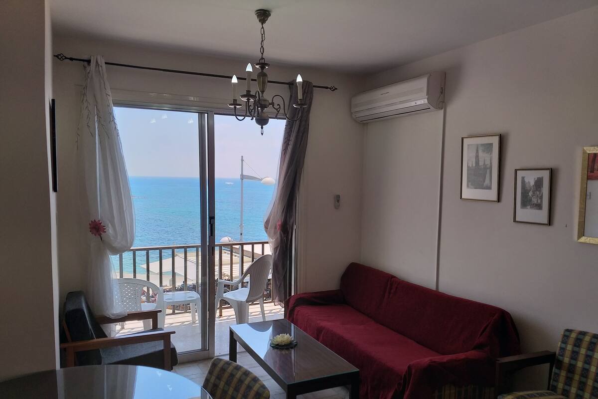 Ein Zimmer mit einem roten Sofa, einem Esstisch mit Stühlen, einer Kristallleuchtre und Blick auf das Meer durch ein großes Fenster.