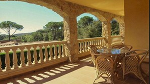Un balcón con arcos de piedra, una mesa de cristal, sillas de mimbre y vistas a árboles y colinas.