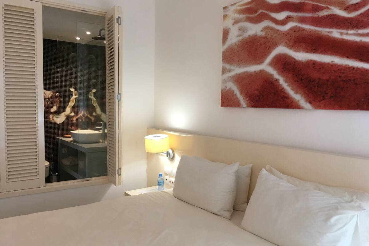 A hotel room with a bed, a nightstand with a lamp, a bathroom with a sink and mirror, and a giraffe-patterned artwork on the wall.