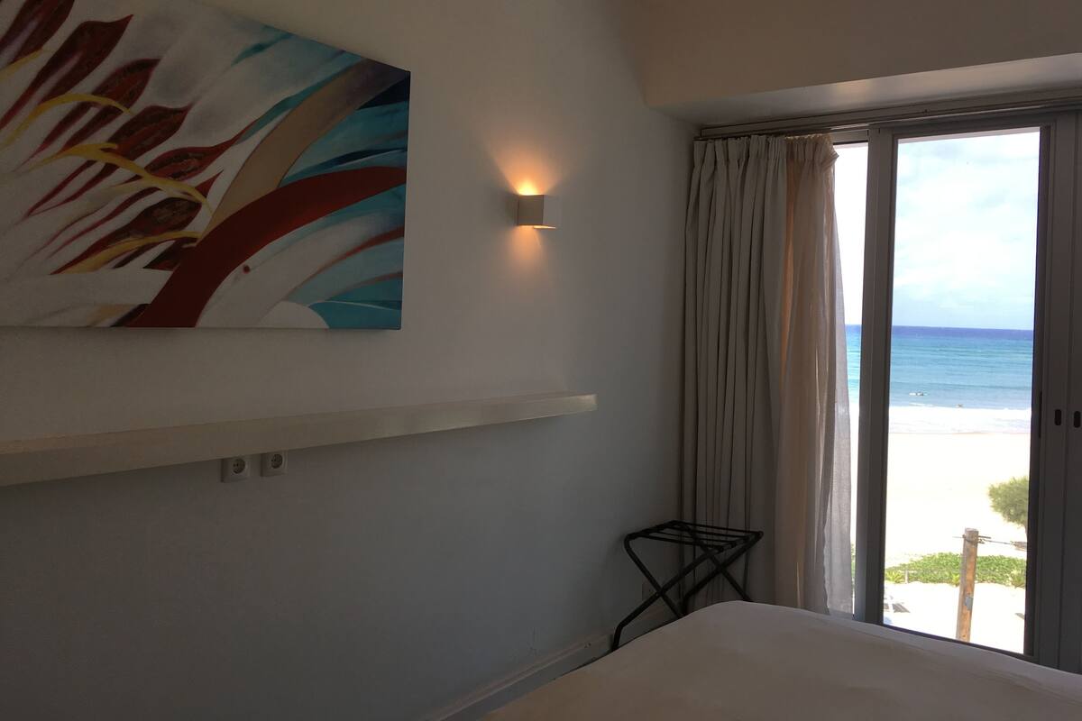 A hotel room with a large window offering a view of the beach, a painting on the wall, and a small black side table.