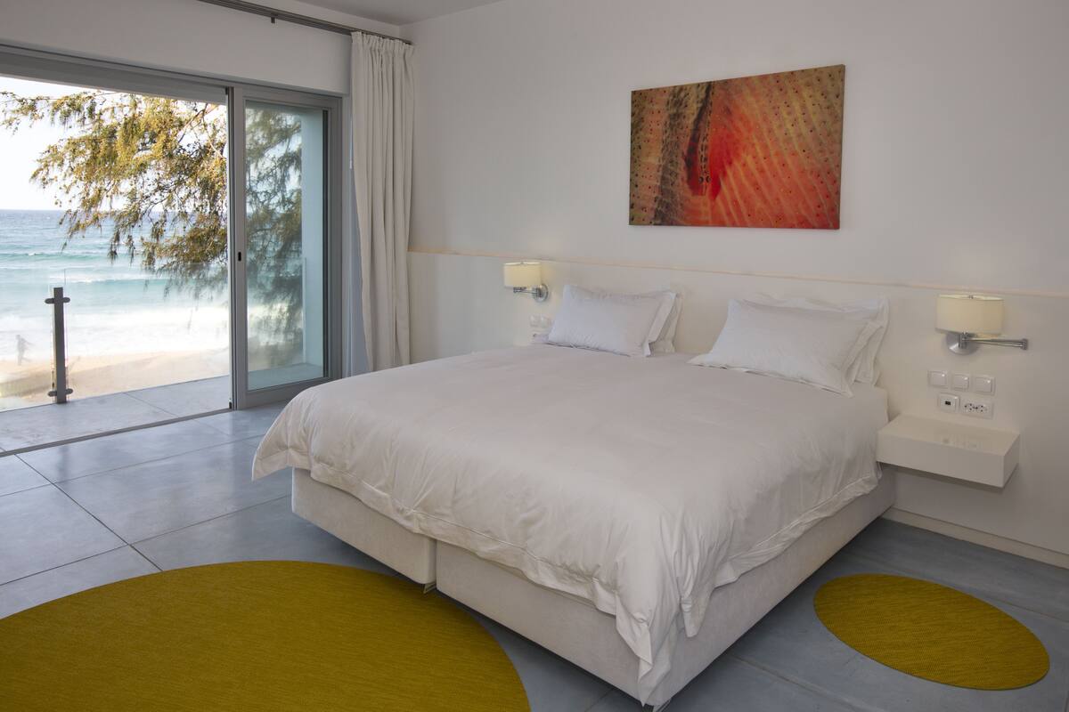 A modern hotel room with a large bed, a sliding glass door to the beach, and a painting on the wall.