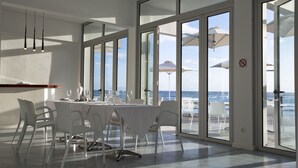 A modern dining area with a large table and chairs, large windows with ocean views, and a ceiling light fixture. - Hotel Tofo Mar (Tofo)