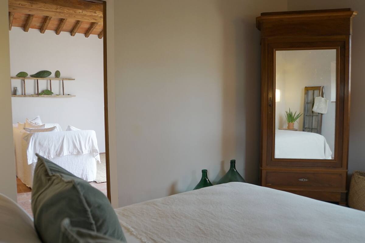 A bedroom with a large wooden wardrobe, a bed with white bedding, and a mirror reflecting a bed and a plant.