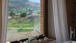 A bathroom with a bathtub, a window with curtains, and a view of a green landscape. - Marrucheti 82 (Campagnatico)