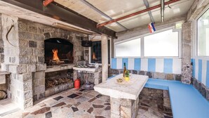 A stone fireplace with a fire, a table with a vase, and a blue bench in a room with a tiled floor and a window with a view.
