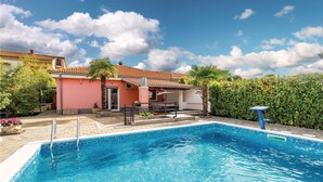 A swimming pool with a tiled edge, a covered patio area, and a pink building with a tiled roof.