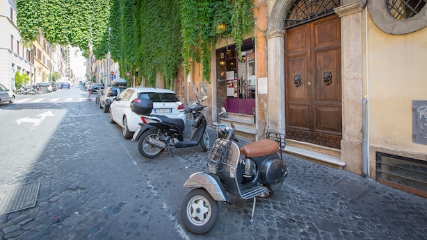 Une rue pavée bordée de voitures et de motos garées, un bâtiment avec une porte en bois et un panneau indiquant une zone de stationnement.