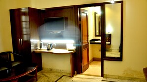 A room with a wooden wardrobe, a small table with a kettle, a chair, and a mirror on the wall. - Hotel Marc Royal (Agra)