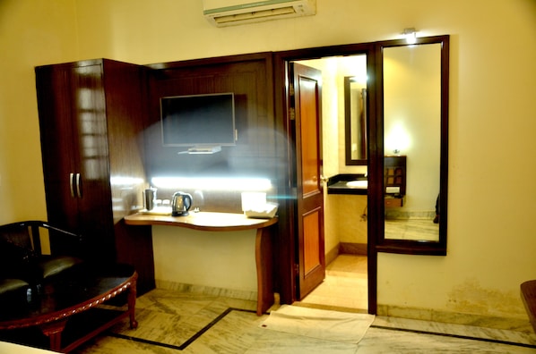 A room with a wooden wardrobe, a small table with a kettle, a chair, and a mirror on the wall. - Hotel Marc Royal (Agra)