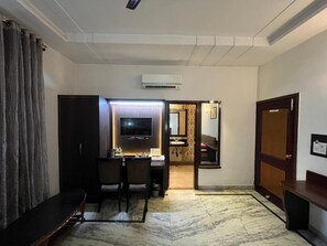A hotel room with a flat-screen TV, a desk with a chair, a sofa, a bathroom with a mirror, and a door. - Hotel Marc Royal (Agra)