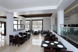 A spacious dining area with multiple tables and chairs, a high ceiling with patterned beams, and large windows with decorative screens. - Hotel Marc Royal (Agra)