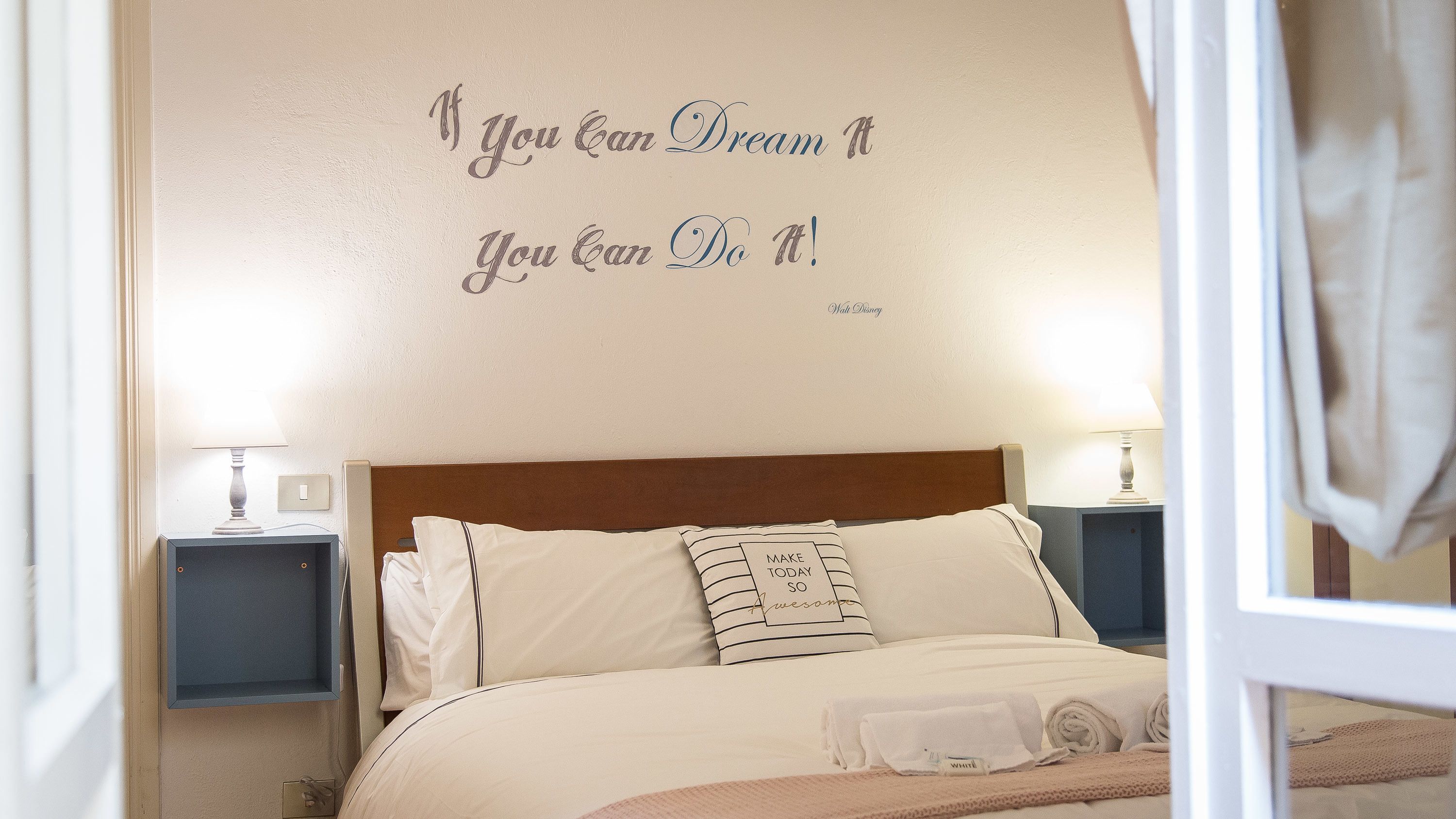 Een net opgemaakt bed met een motiverende quote op de muur, een bedlampje en een nachtkastje.