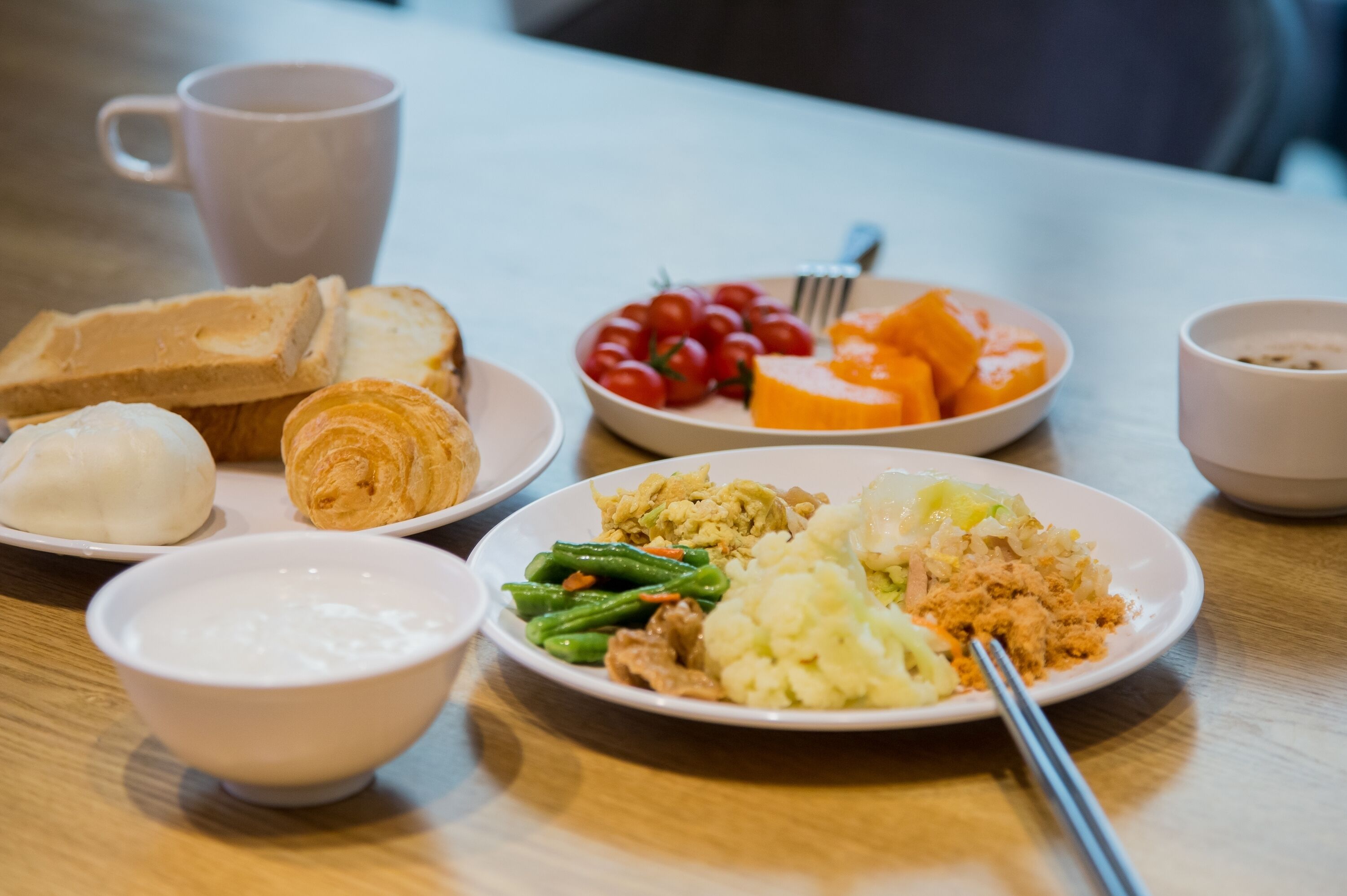 A table with various breakfast items including toast, croissants, a bowl of soup, a plate with tomatoes and cheese, and a cup of coffee.