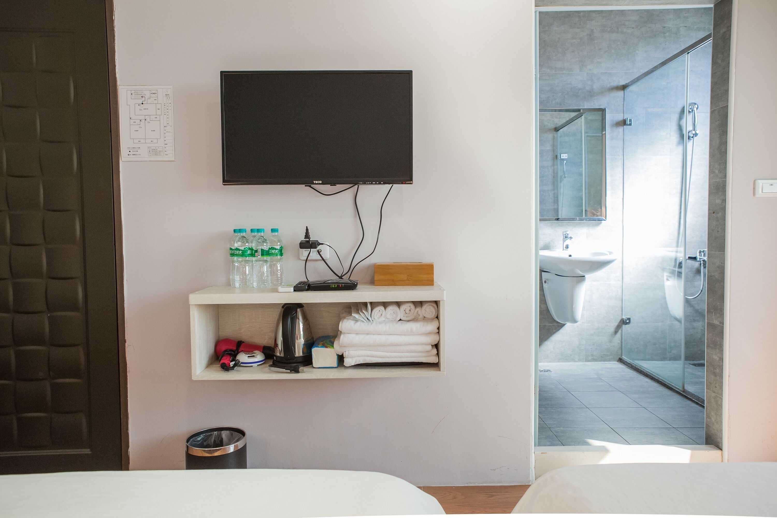 A hotel room with a TV mounted on the wall, a shelf with toiletries, and a view into the bathroom.