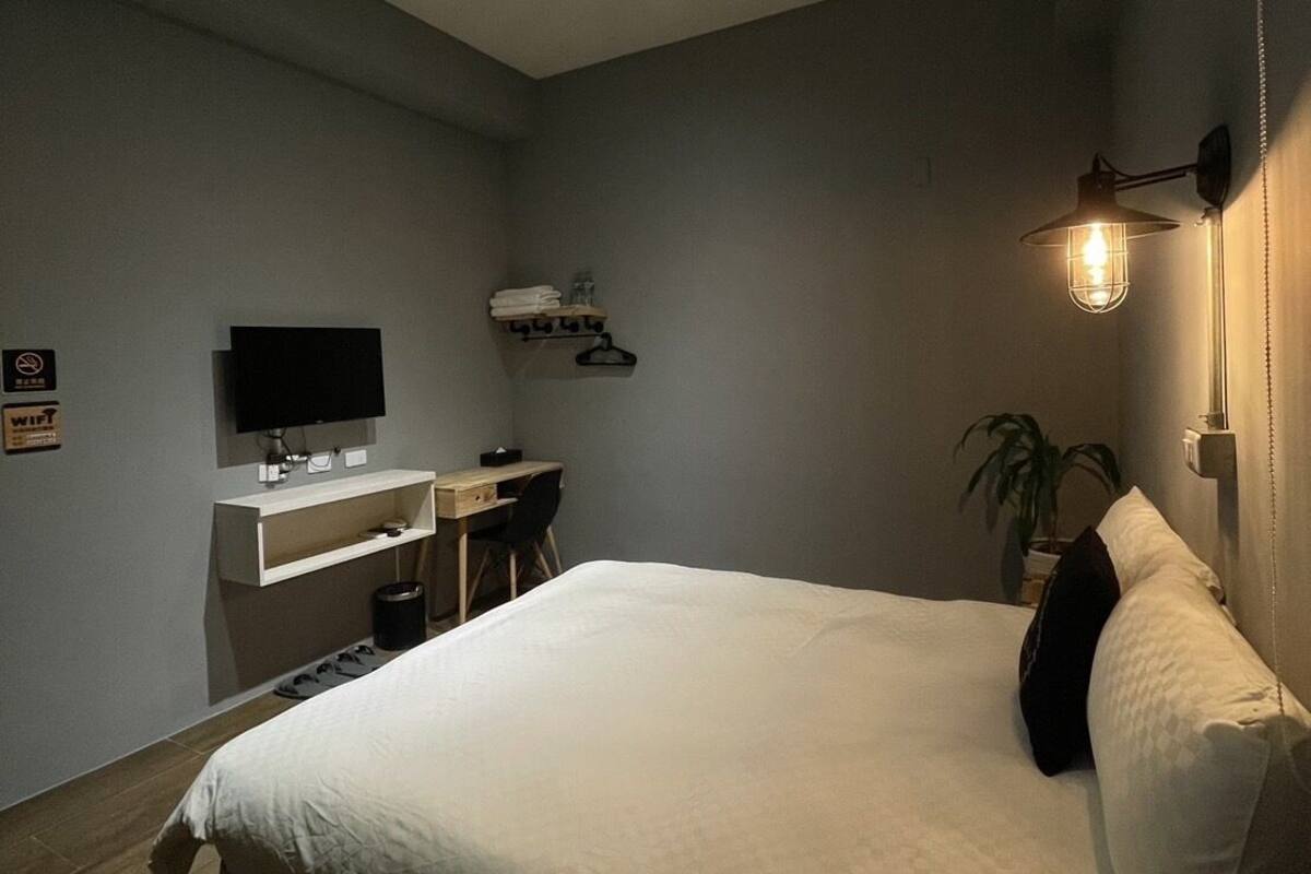 A modern hotel room with a large bed, a desk with a chair, a flat-screen TV, and a wall-mounted lamp.