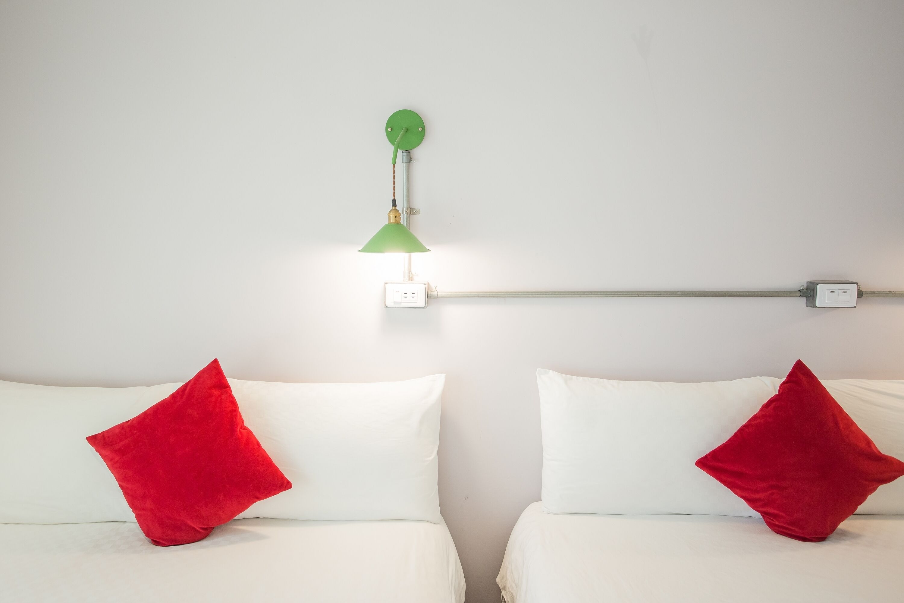 A room with two beds, each with a white mattress and a red pillow. A green wall-mounted lamp is visible above the beds.