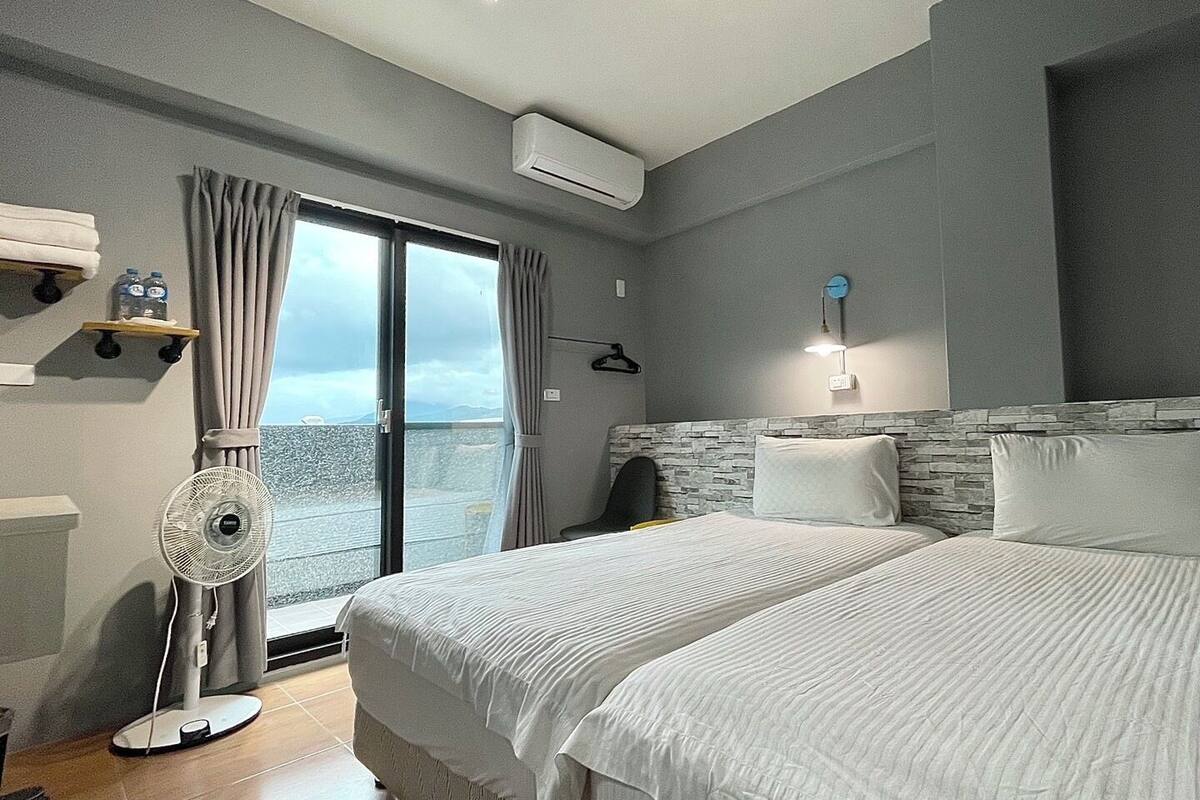 A hotel room with two beds, a fan, a window with curtains, a wall-mounted heater, and a view of the sea.