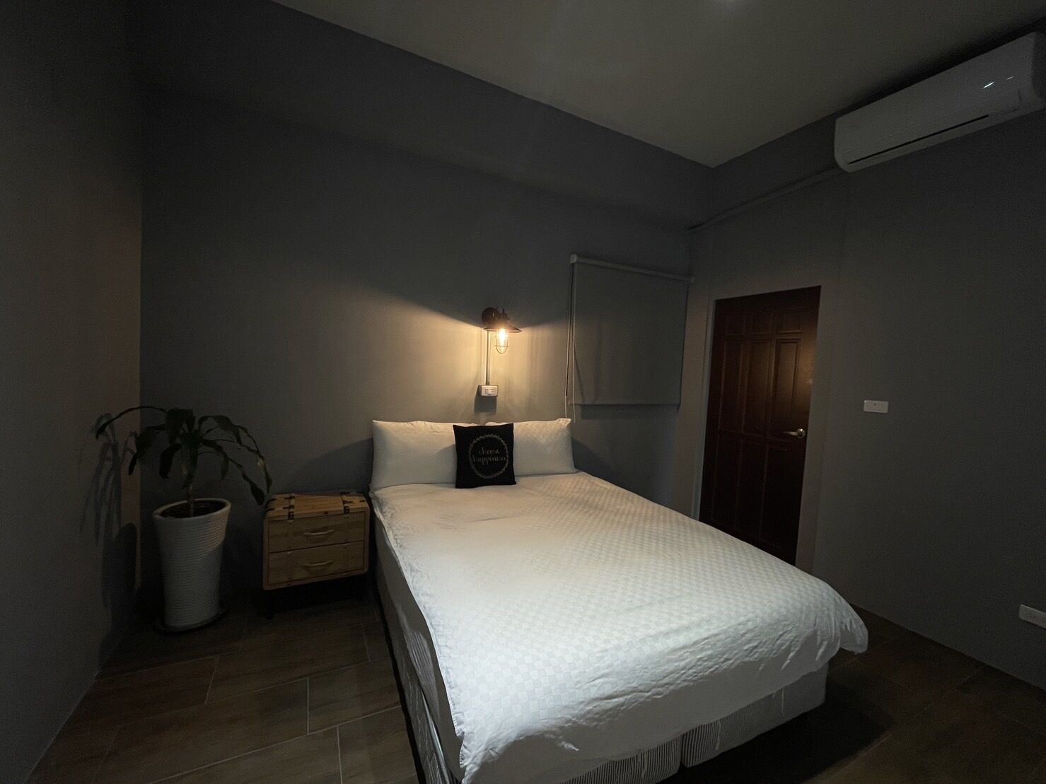 A bedroom with a bed, bedside table, lamp, potted plant, and a door.