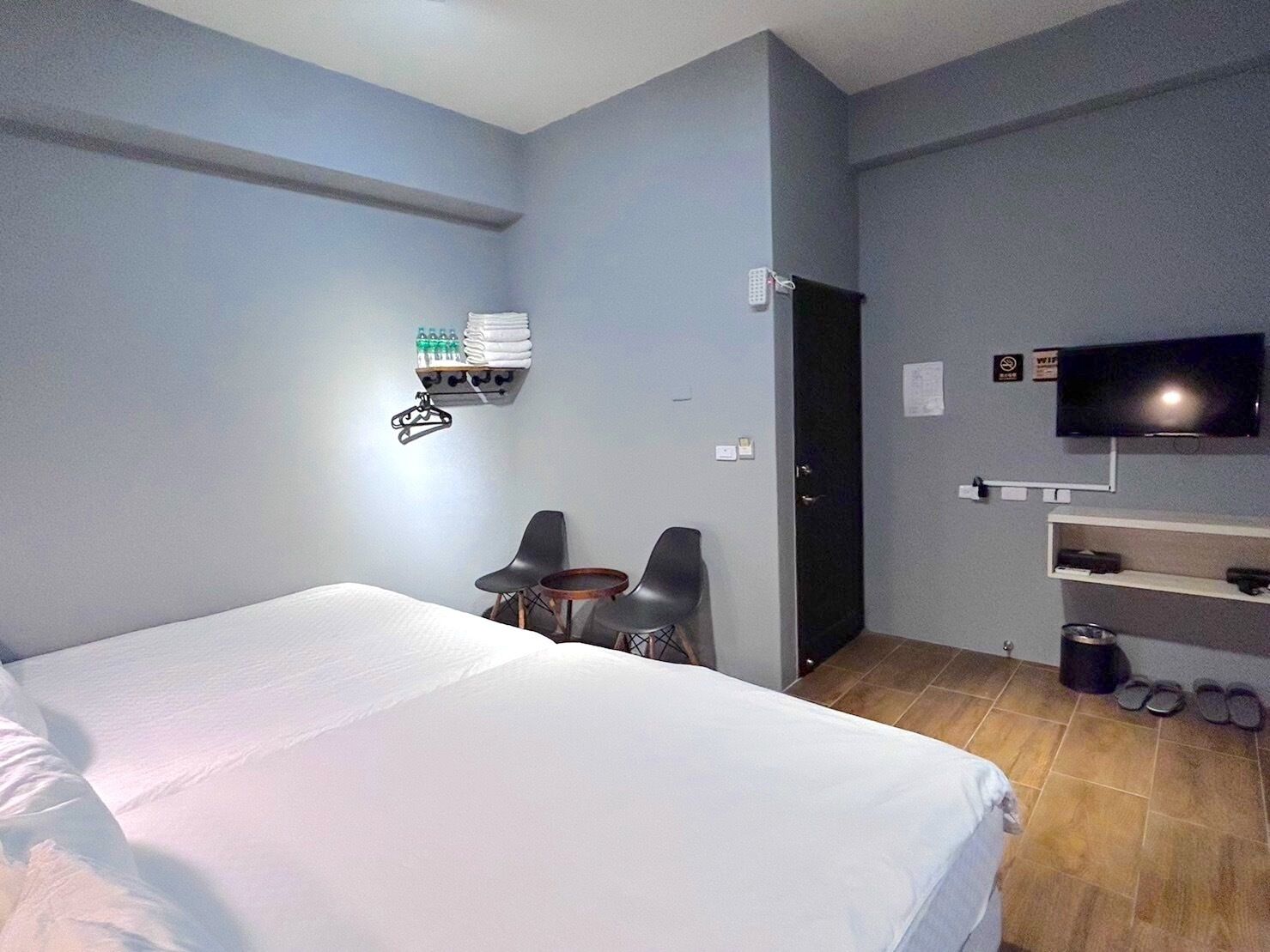 Une chambre d’hôtel avec un lit, deux chaises, une télévision, une étagère avec des articles de toilette et une porte.