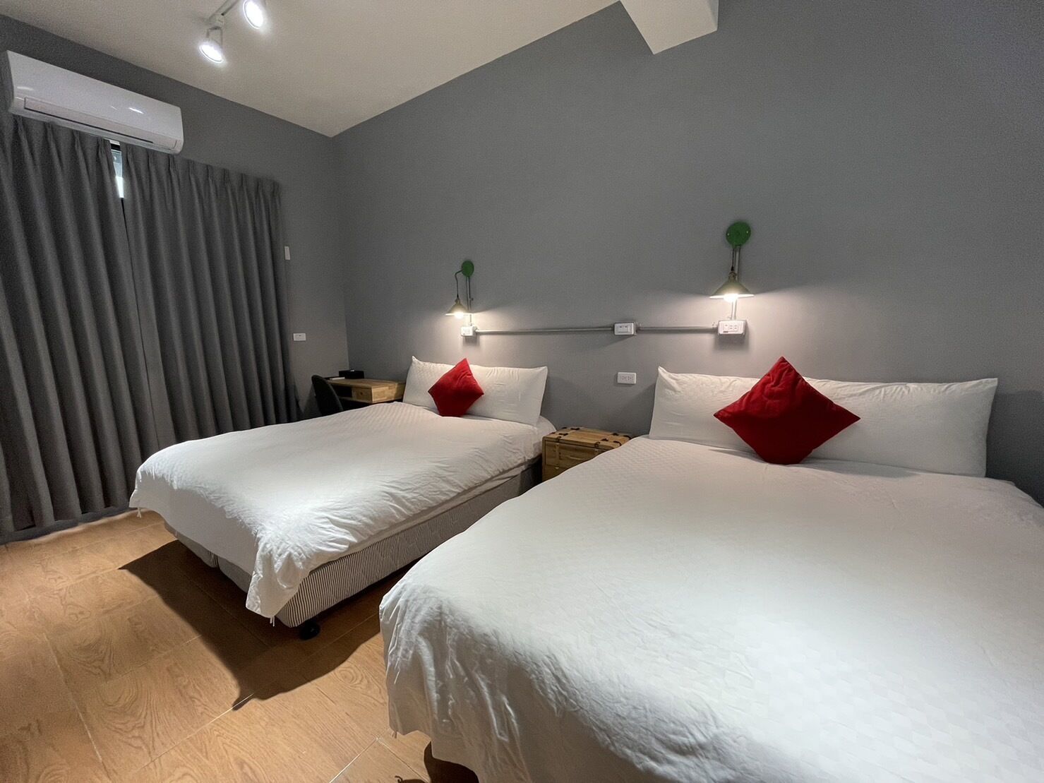 Habitación de hotel con dos camas, cortinas grises, suelo de madera y aire acondicionado instalado en la pared.