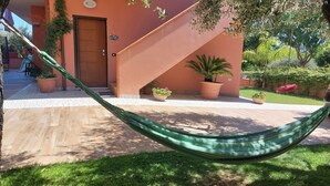 Una hamaca en un espacio exterior soleado con una escalera, plantas en macetas y un tobogán al fondo.