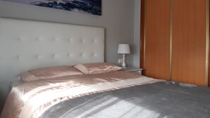 A bedroom with a large bed, a nightstand with a lamp, a painting of a lighthouse, and a wooden wardrobe.